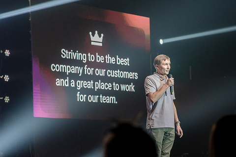 A speaker presenting on stage, beside a screen with a message on creating a great workplace.